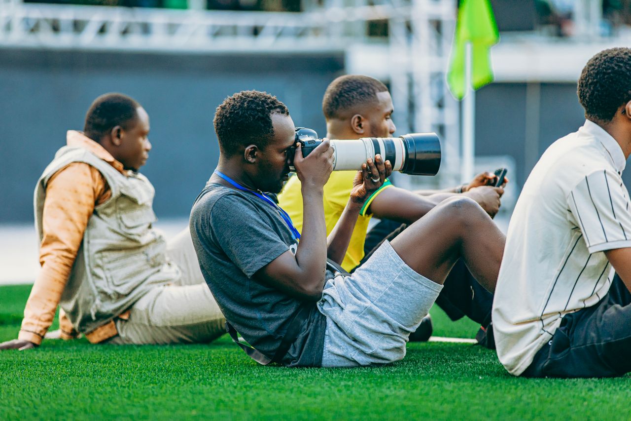 heros-img Group of young photographers sitting on grass, capturing the scene with professional cameras.